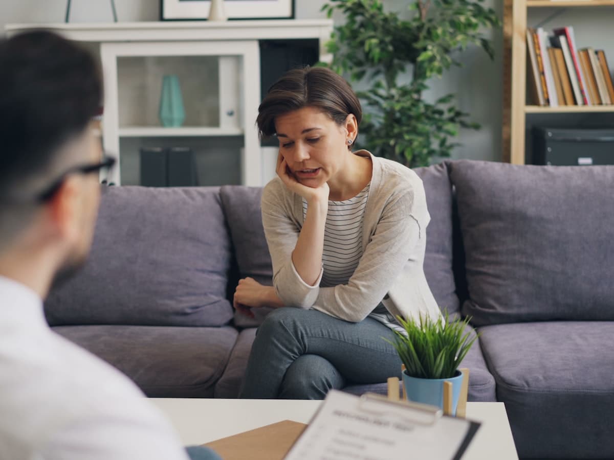 Two people talking in a bright room with a sofa and a plant